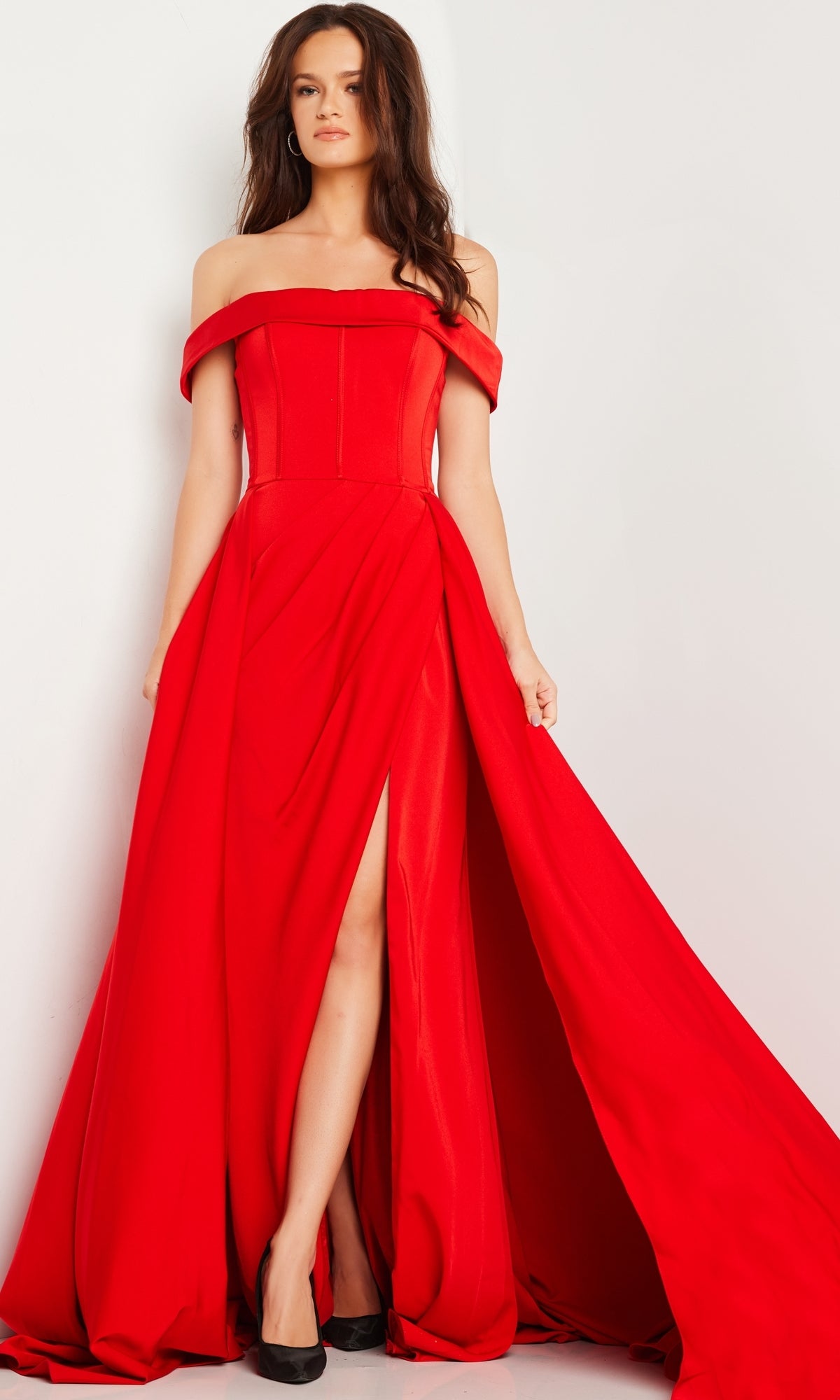 A woman stands against a white background wearing the Jovani Off-Shoulder Long A-Line Prom Dress 09874, a floor-length red gown with a thigh-high slit.